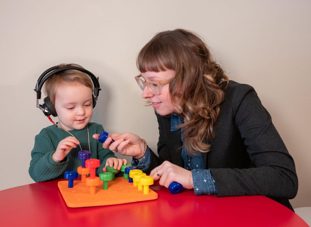 Child wearing headphones and playing with colorful stacking toys, assisted by an adult, in a learning environment.