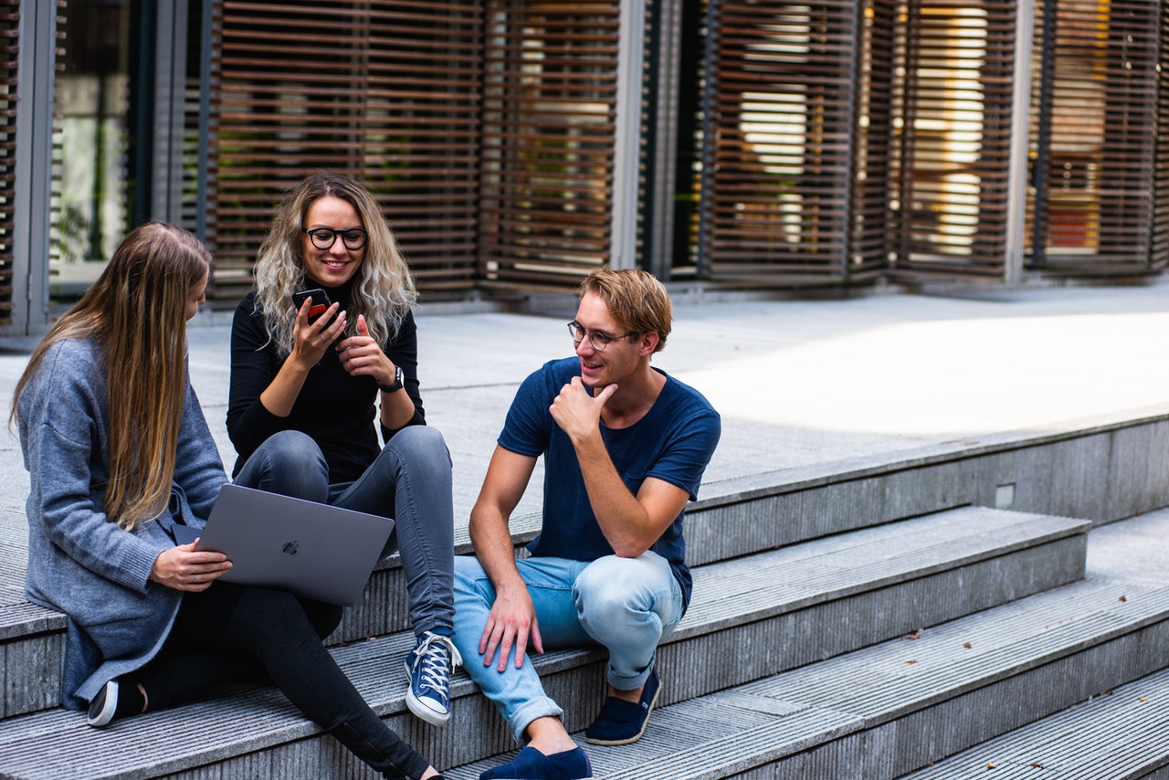 Three young adults engaged in conversation on outdoor steps, one using a laptop, with a focus on collaboration and technology in a modern setting.